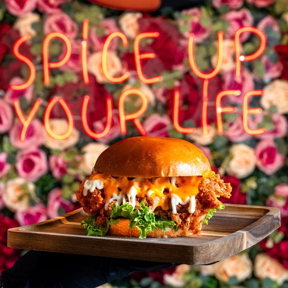 A Tikkaville fried chicken burger with sauces and lettuce on a wooden tray, in front of a floral wall with a neon sign.