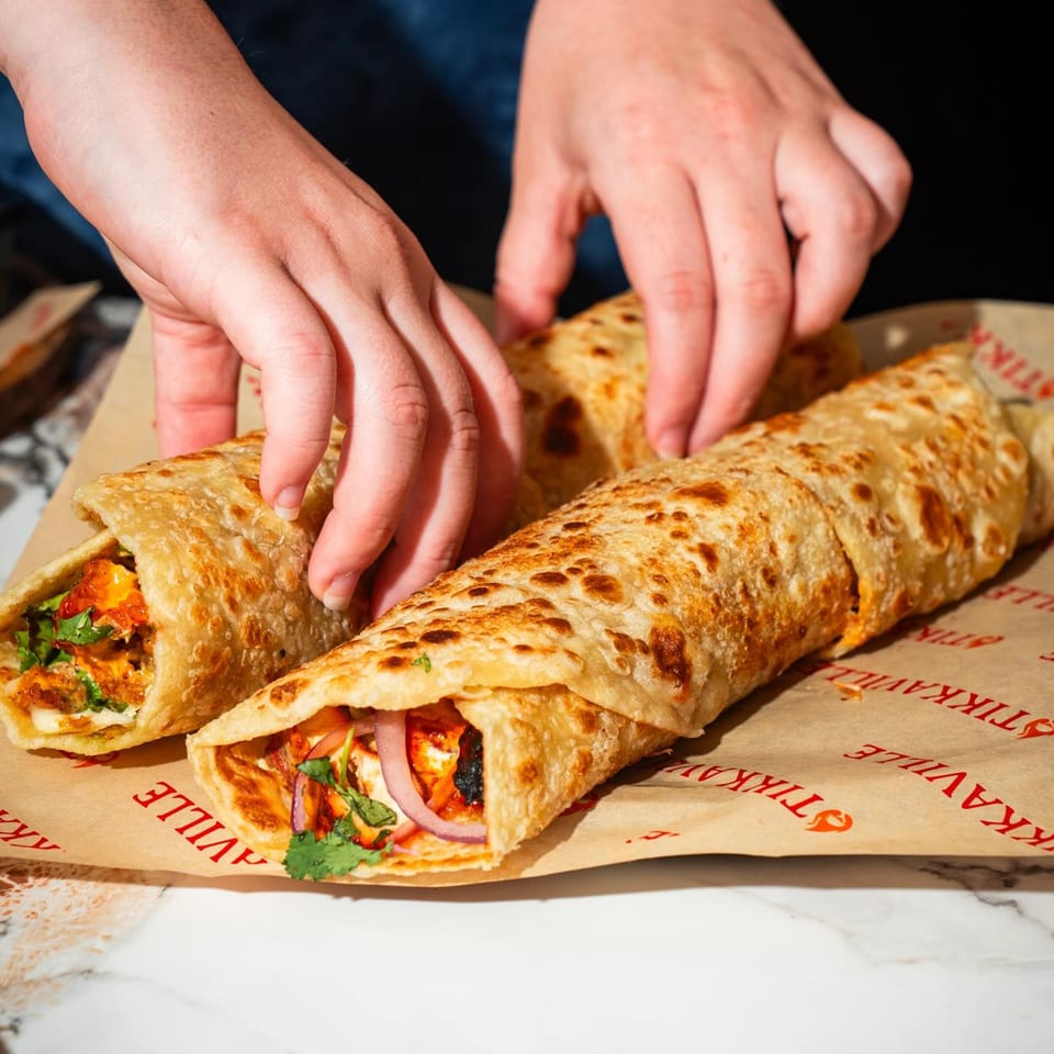 Hands arranging two Tikkaville chicken tikka rolls with red onions and cilantro on branded paper.