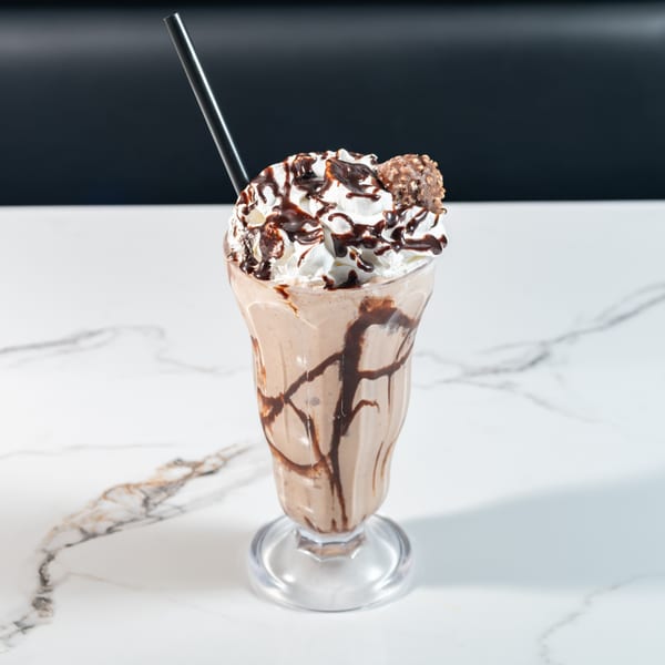 Chocolate milkshake with whipped cream, chocolate drizzle, and a chocolate candy on a marble table.