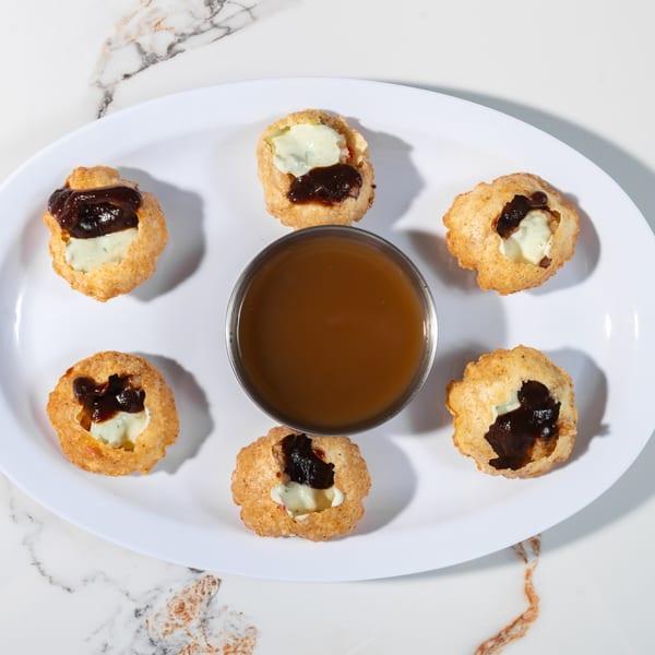 Six pani puri shells filled with white and dark sauces, surrounding a bowl of brown liquid.