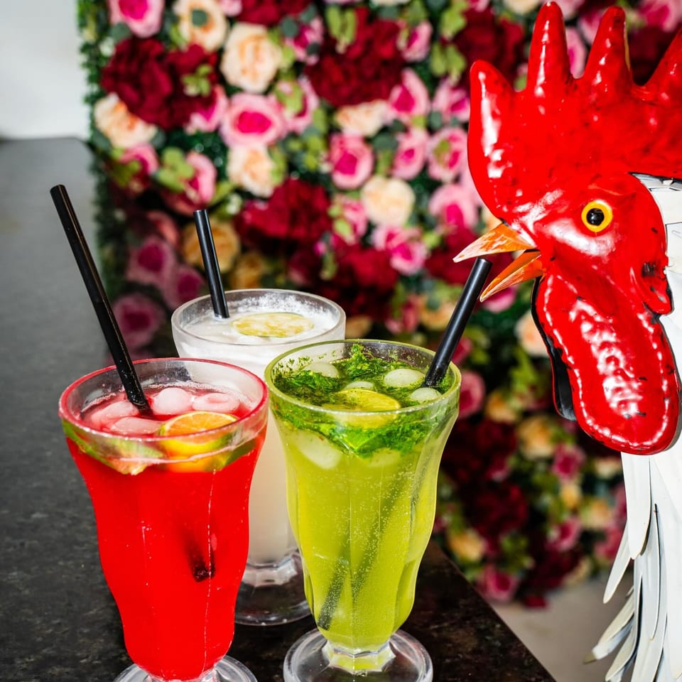 Three colorful Tikkaville drinks with straws, garnished with fruit and mint, next to a rooster statue.