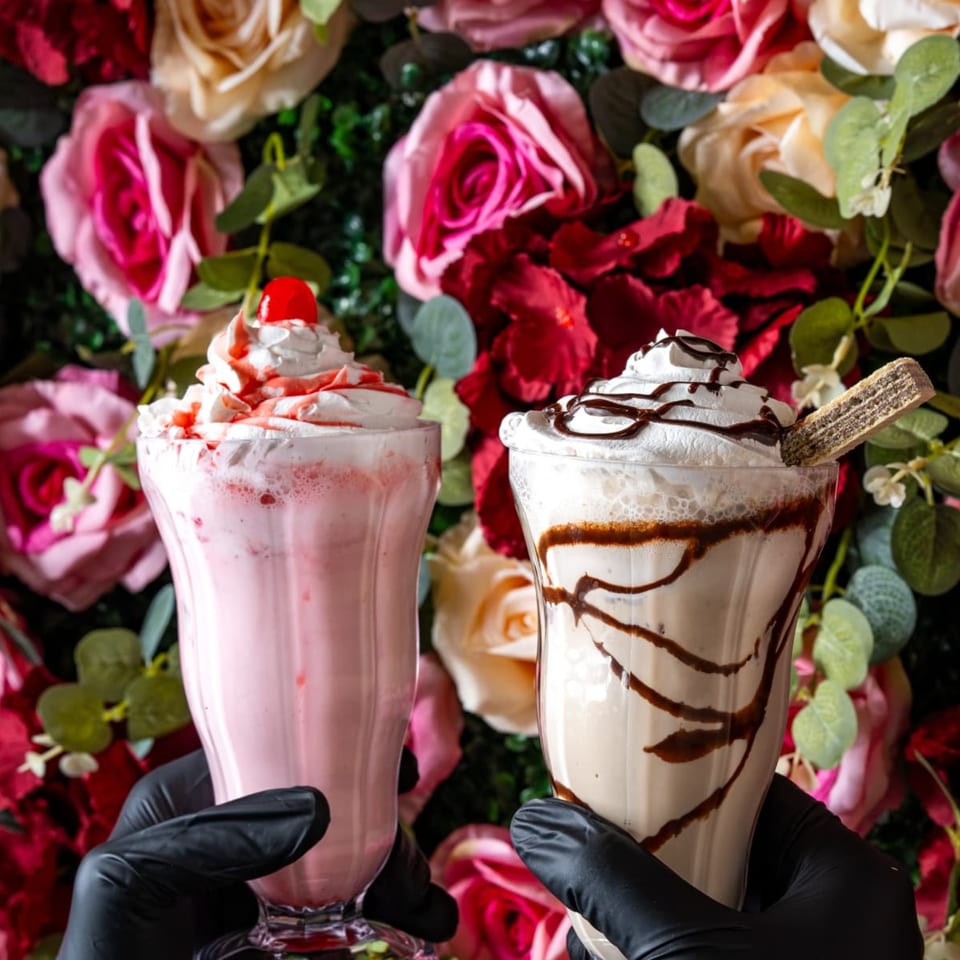 Two hands in black gloves holding a strawberry milkshake and a chocolate milkshake against a floral background.